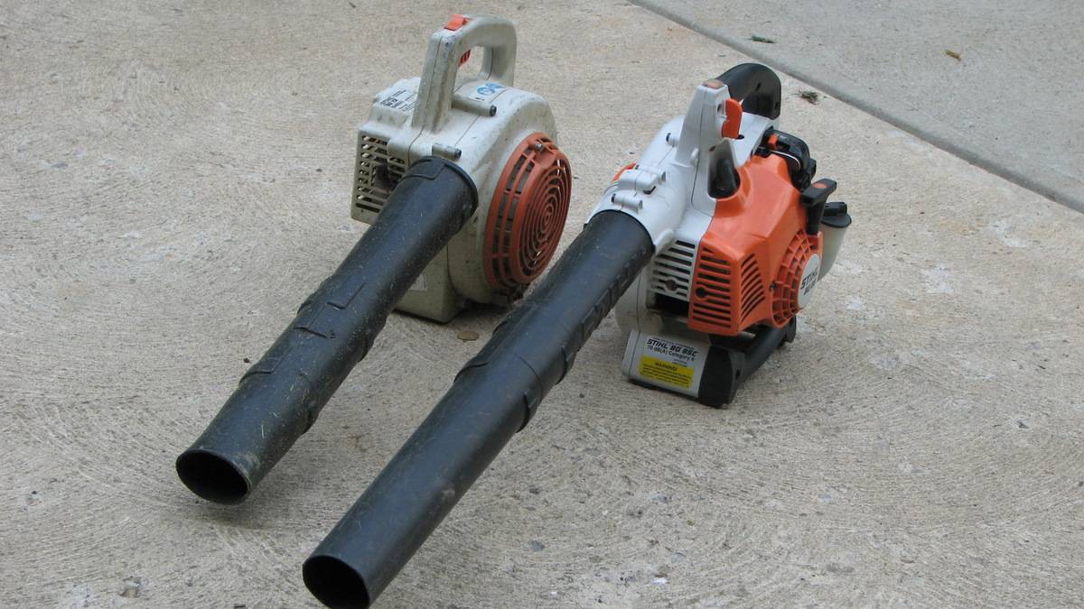 Two handheld leaf blowers with long black nozzles resting on a concrete surface.”