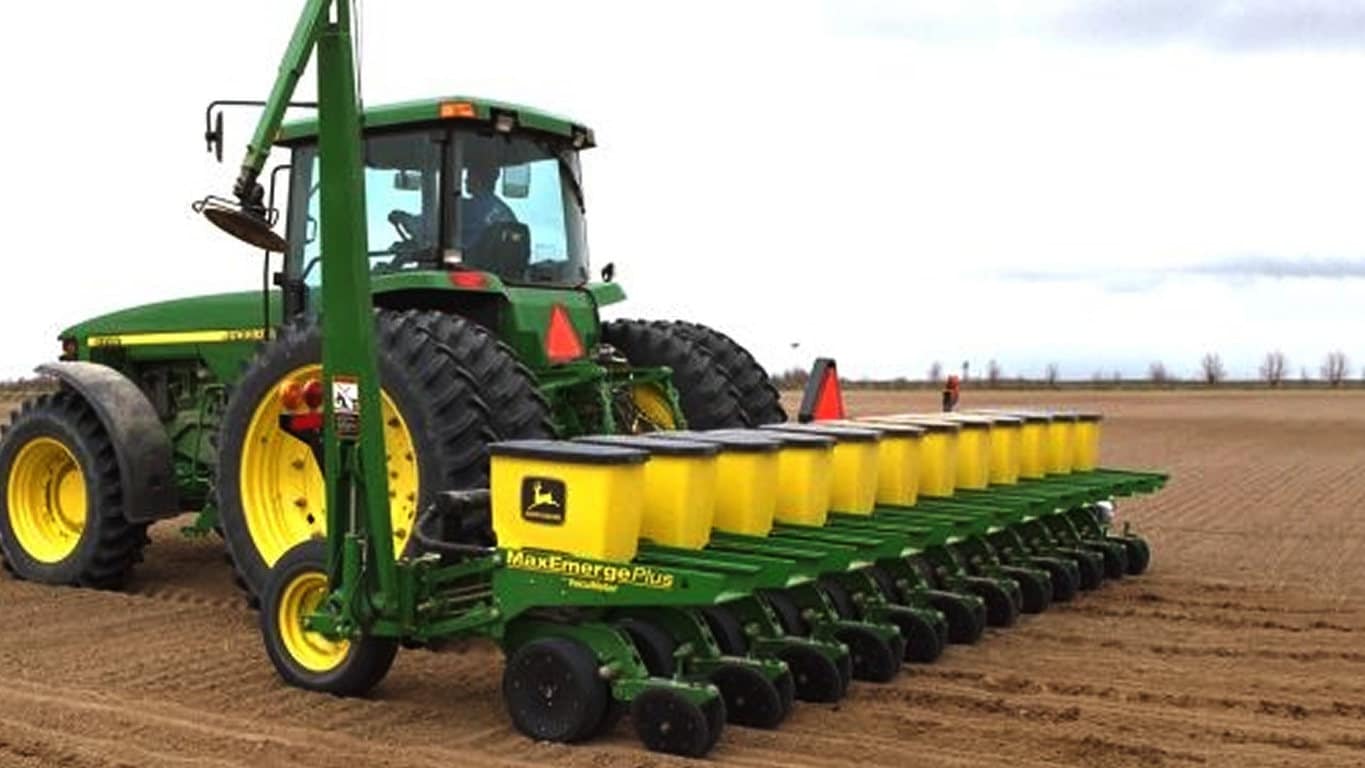 A green tractor pulling a multi-row seed planter across a plowed agricultural field.