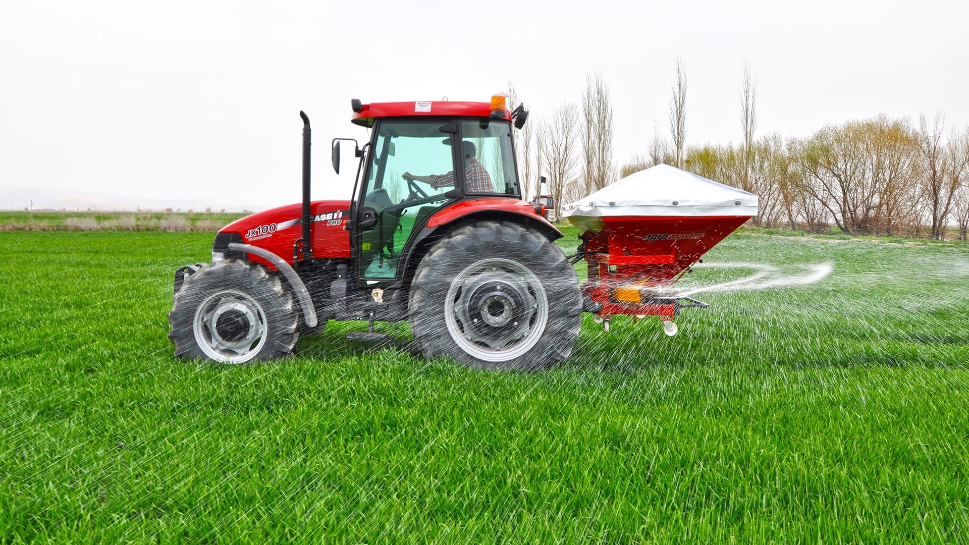 Tractor-mounted broadcast fertilizer spreader applying fertilizer across a green field.